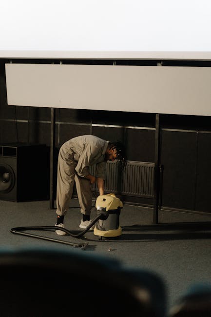 A person kneeling on a patterned carpet in a residential interior, using a yellow and black portable vacuum cleaner equipped with a hose to perform surface cleaning. The individual is wearing a khaki-