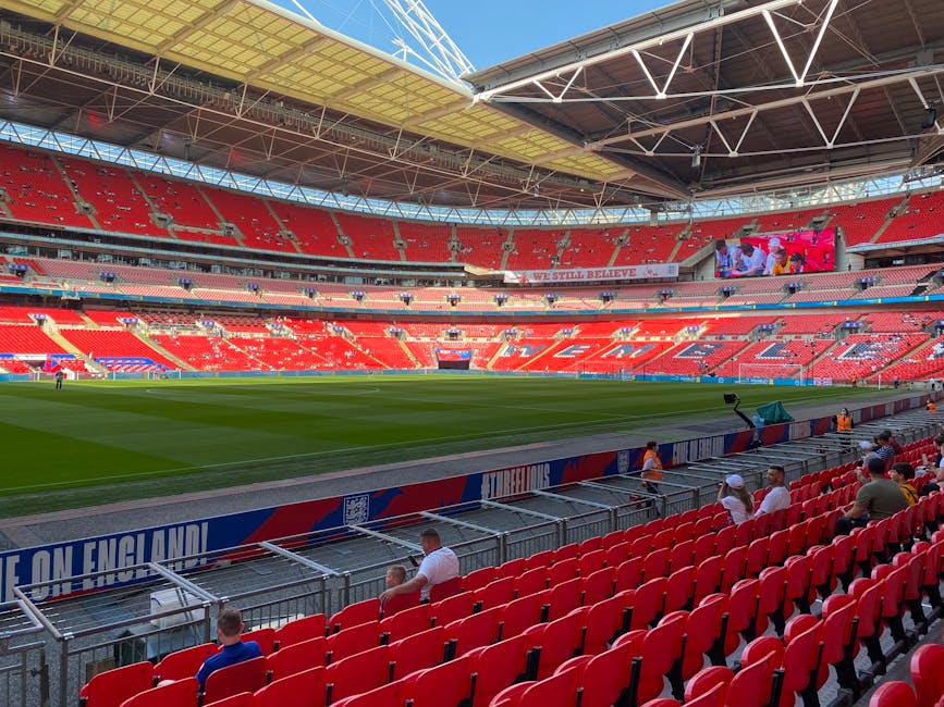 Interior view of Wembley Stadium, featuring a well-maintained grass football pitch with bright green, evenly trimmed surface and clearly marked white lines, surrounded by red seating sections and part